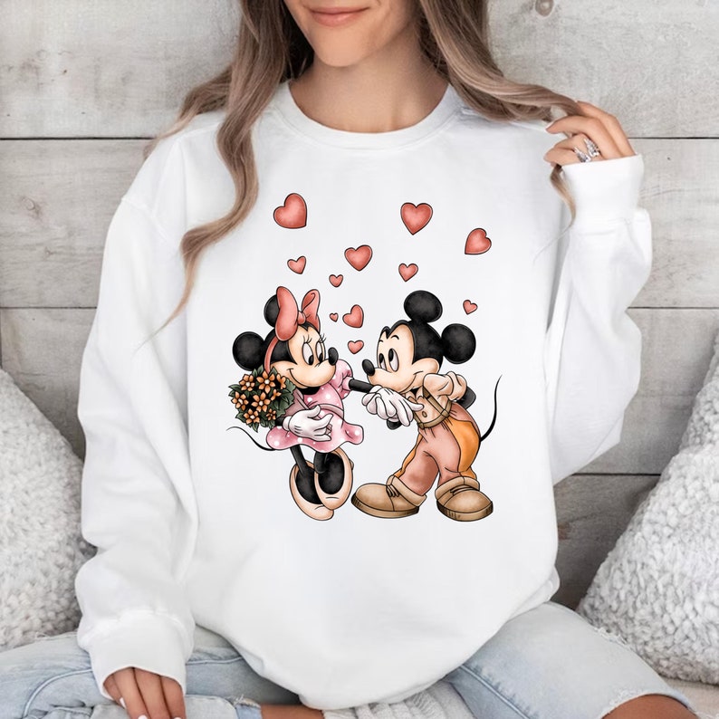 May include: White sweatshirt with a graphic print of Mickey Mouse and Minnie Mouse holding hands with hearts floating around them.