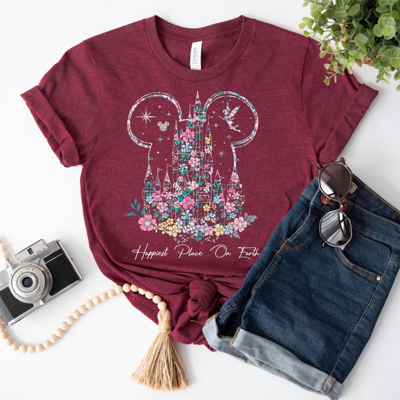 May include: A maroon t-shirt with a floral design of a castle in the shape of Mickey Mouse ears. The text "Happiest Place On Earth" is printed below the castle.