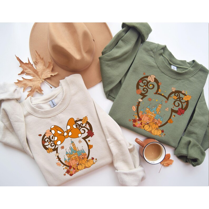 May include: Two sweatshirts with a fall-themed Mickey Mouse design. One sweatshirt is beige with a bow and the other is olive green with Mickey Mouse ears.