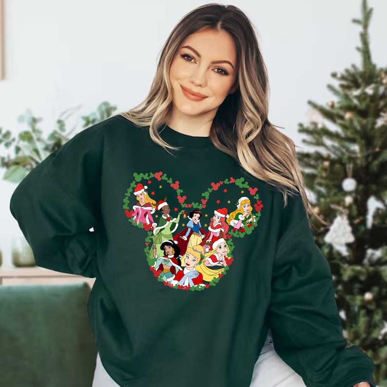 Pu&ograve; includere: Felpa verde scuro con un design a forma di testa di Topolino riempita di principesse Disney con cappelli da Babbo Natale. Le principesse sono circondate da bacche rosse e foglie verdi, creando un tema festivo natalizio.