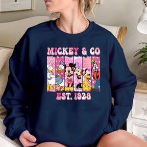 Puede incluir: Sudadera azul marino con un gr&aacute;fico rosa y blanco de Mickey Mouse y sus amigos. El gr&aacute;fico incluye el texto "Mickey & Co. Est. 1928".