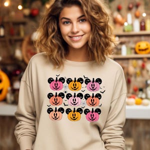 May include: A beige sweatshirt with a pattern of orange, pink, and peach Mickey Mouse pumpkins. The pumpkins have black eyes, noses, and mouths. There are also black bats and spiderwebs on the sweatshirt.