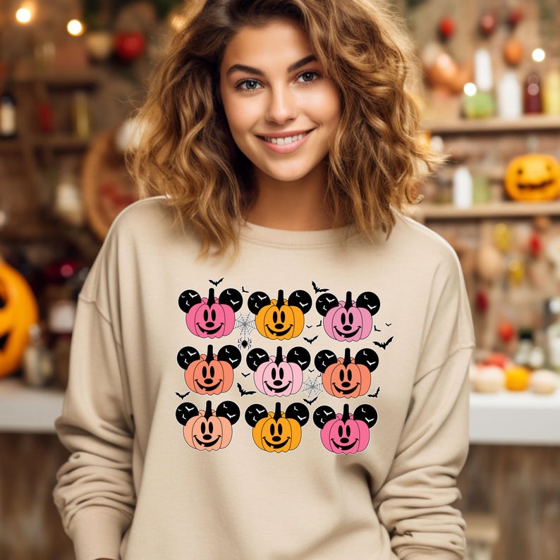 May include: A beige sweatshirt with a pattern of orange, pink, and peach Mickey Mouse pumpkins. The pumpkins have black eyes, noses, and mouths. There are also black bats and spiderwebs on the sweatshirt.