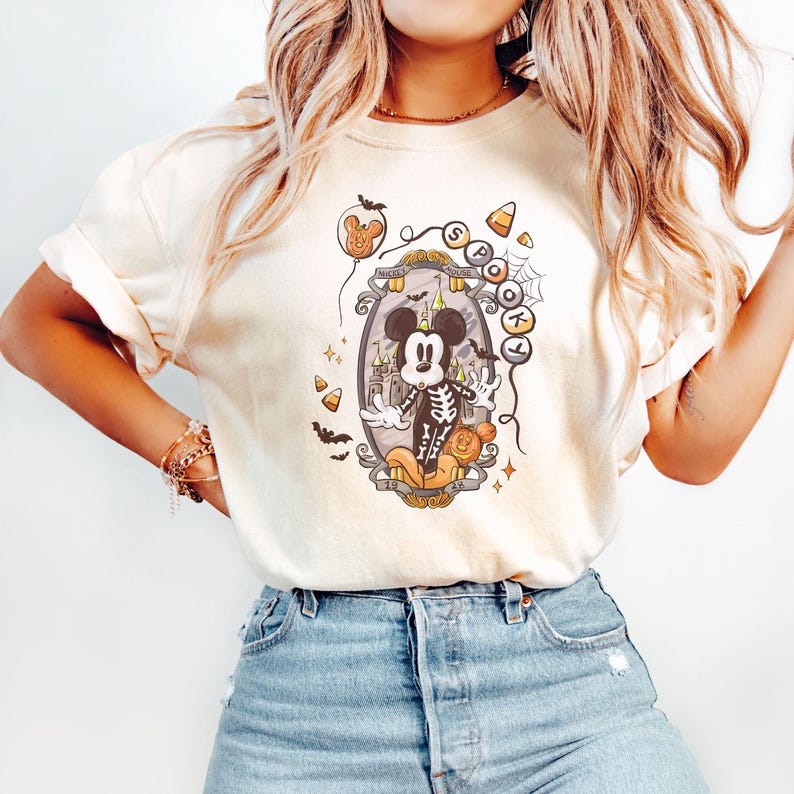 May include: Cream-colored t-shirt featuring a Halloween-themed graphic. The design includes a cartoon Mickey Mouse dressed as a skeleton, surrounded by Halloween elements like candy corn, bats, and a haunted house. The text "Spooky" is also visible.