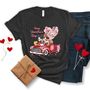 May include: A dark grey t-shirt with a graphic of Mickey Mouse and Minnie Mouse in a red truck with pink heart balloons. The text "Happy Valentine's Day" is printed on the shirt.