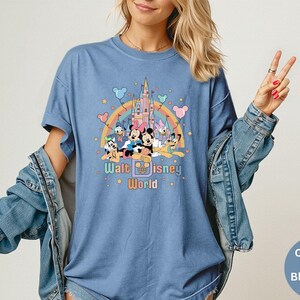 Puede incluir: Camiseta azul vaquero con un gr&aacute;fico colorido de personajes de Disney, un arco&iacute;ris y un castillo. El texto "Walt Disney World" est&aacute; impreso debajo. La camiseta tiene mangas cortas y un corte holgado.