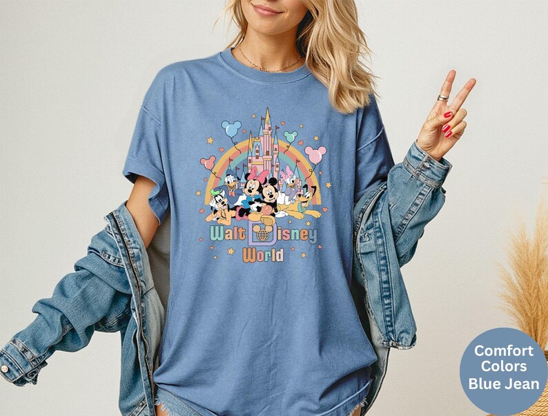 Puede incluir: Camiseta azul vaquero con un gr&aacute;fico colorido de personajes de Disney, un arco&iacute;ris y un castillo. El texto "Walt Disney World" est&aacute; impreso debajo. La camiseta tiene mangas cortas y un corte holgado.