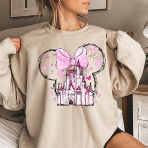 May include: A beige sweatshirt with a pink bow and a pink castle graphic. The castle is surrounded by hearts and stars.