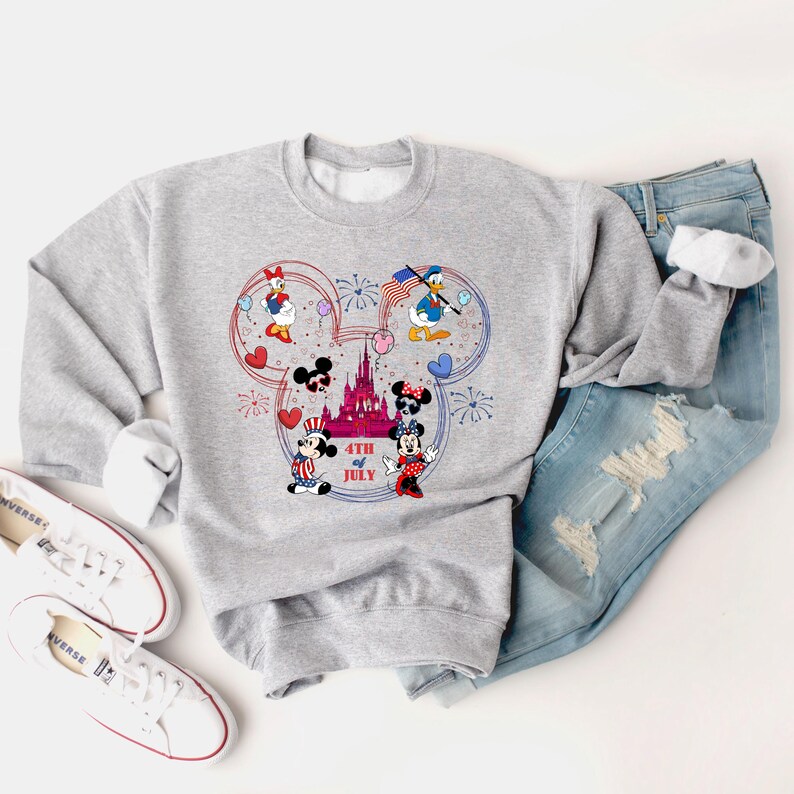 Peut inclure: Un sweat-shirt gris clair avec un motif Mickey Mouse sur le th&egrave;me du 4 juillet. Le graphisme comprend Mickey, Minnie, Donald et Daisy, avec des feux d'artifice et un ch&acirc;teau. Le texte "4th of July" est &eacute;galement inclus.