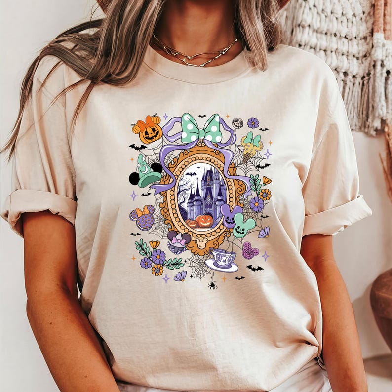 Puede incluir: Camiseta beige con un dise&ntilde;o de Halloween. El dise&ntilde;o incluye un castillo, calabazas y personajes de dibujos animados. El gr&aacute;fico est&aacute; enmarcado por un borde dorado y adornado con un lazo verde menta y detalles morados.