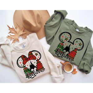 Puede incluir: Dos sudaderas de color crema con un dise&ntilde;o de Mickey Mouse con tem&aacute;tica navide&ntilde;a. El dise&ntilde;o presenta un lazo de cuadros rojos y verdes, una silueta del castillo de Cenicienta y la palabra "Believe".