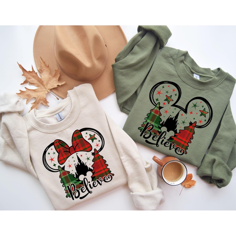 Puede incluir: Dos sudaderas de color crema con un dise&ntilde;o de Mickey Mouse con tem&aacute;tica navide&ntilde;a. El dise&ntilde;o presenta un lazo de cuadros rojos y verdes, una silueta del castillo de Cenicienta y la palabra "Believe".