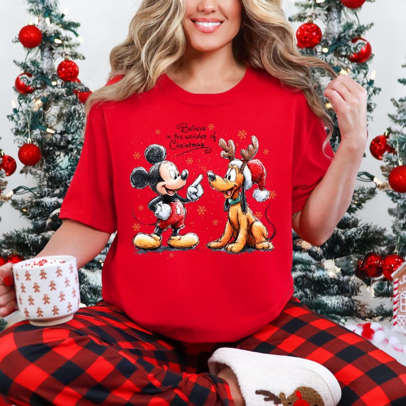 May include: Red t-shirt featuring Mickey Mouse and Pluto in Christmas attire. Mickey Mouse is wearing a red and white outfit, pointing at Pluto, who is wearing a Santa hat and reindeer antlers. The text "Believe in the wonder of Christmas" is above the characters.