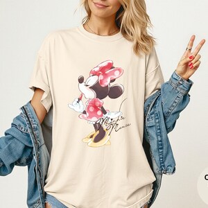 K&ouml;nnte beinhalten: Elfenbeinfarbenes T-Shirt mit einem Minnie Mouse-Motiv. Die Zeichentrickfigur ist in einem roten gepunkteten Kleid und einer Schleife mit gelben Schuhen dargestellt. Die Worte "Minnie Mouse" sind unter dem Bild in Kursivschrift geschrieben. Das Shirt ist Teil der "Comfort Colors"-Kollektion.