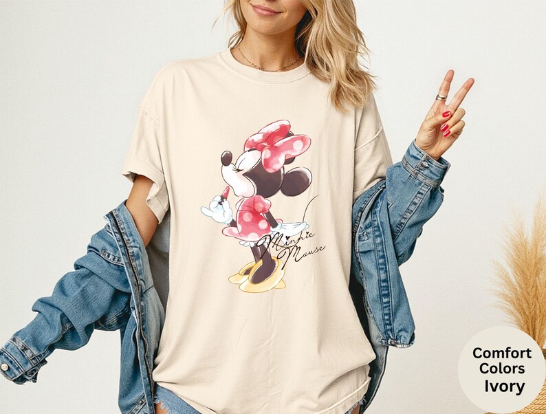 K&ouml;nnte beinhalten: Elfenbeinfarbenes T-Shirt mit einem Minnie Mouse-Motiv. Die Zeichentrickfigur ist in einem roten gepunkteten Kleid und einer Schleife mit gelben Schuhen dargestellt. Die Worte "Minnie Mouse" sind unter dem Bild in Kursivschrift geschrieben. Das Shirt ist Teil der "Comfort Colors"-Kollektion.