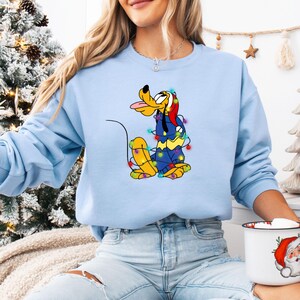K&ouml;nnte beinhalten: Hellblaues Sweatshirt mit einem Cartoon-Pluto-Charakter, der mit bunten Weihnachtsbeleuchtungen geschm&uuml;ckt ist. Pluto zeigt seine Zunge und tr&auml;gt einen Weihnachtsmannhut. Das Sweatshirt ist einfarbig hellblau, passend zur Weihnachtszeit.