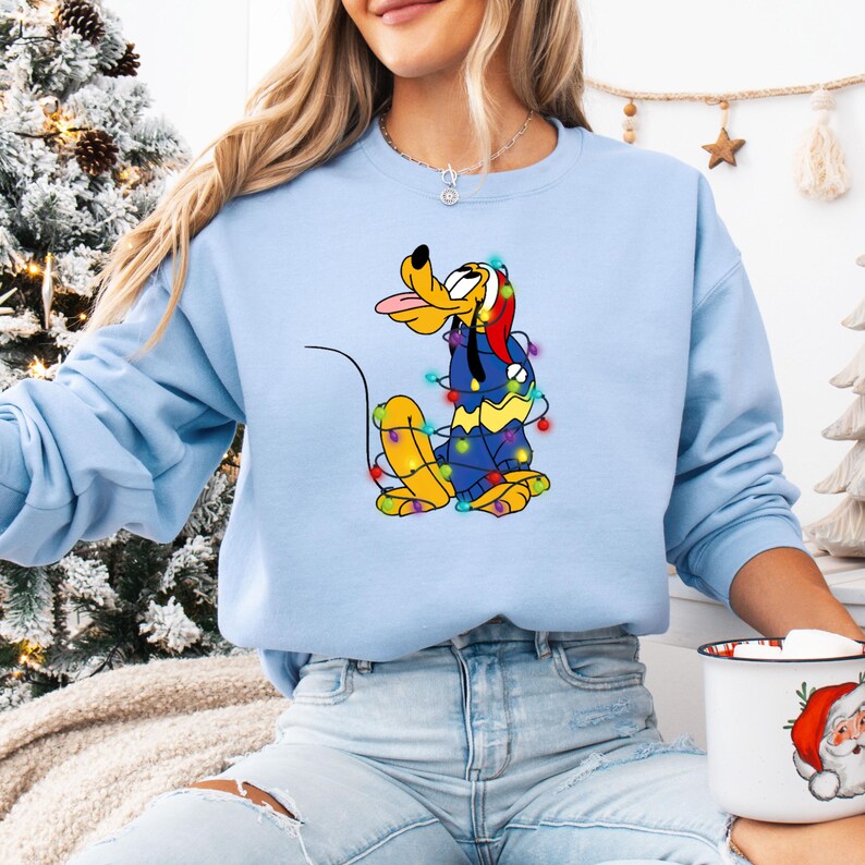 K&ouml;nnte beinhalten: Hellblaues Sweatshirt mit einem Cartoon-Pluto-Charakter, der mit bunten Weihnachtsbeleuchtungen geschm&uuml;ckt ist. Pluto zeigt seine Zunge und tr&auml;gt einen Weihnachtsmannhut. Das Sweatshirt ist einfarbig hellblau, passend zur Weihnachtszeit.