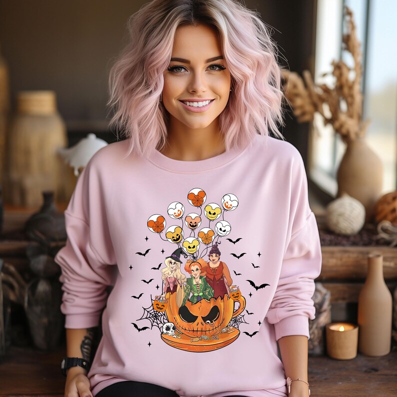 May include: Pink sweatshirt with a graphic of three witches flying in a pumpkin-shaped mug with Mickey Mouse-shaped balloons. The witches are wearing Halloween costumes and the mug has a spooky face.
