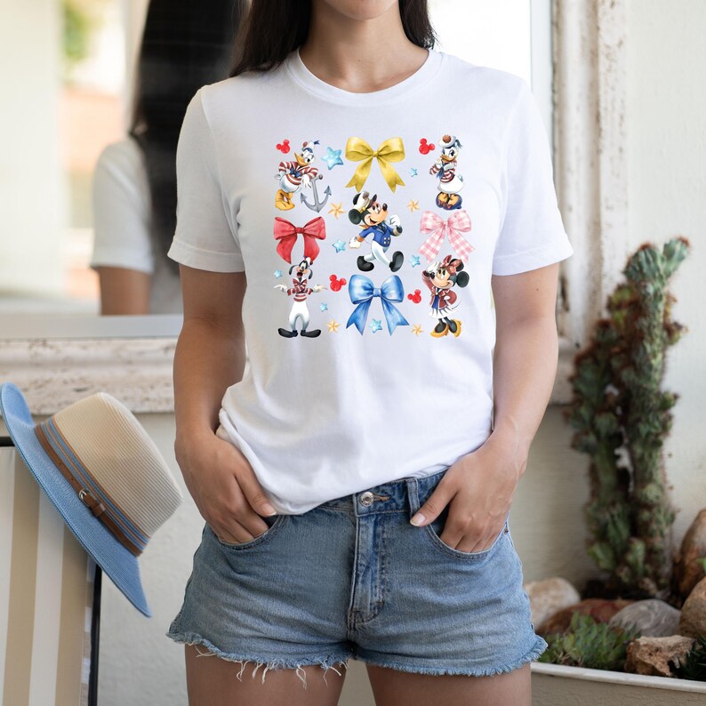 May include: White t-shirt with a colorful print of Mickey Mouse and friends wearing sailor outfits and holding red, blue, and yellow bows. The print also includes stars and anchors.