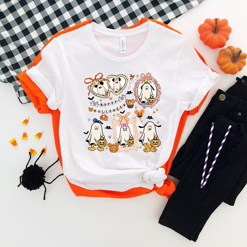 Puede incluir: Camiseta blanca con un dise&ntilde;o de Halloween con fantasmas de dibujos animados, calabazas y el texto "HAPPY HALLOWEEN". La camiseta se combina con vaqueros negros, calabazas naranjas y decoraciones de Halloween.