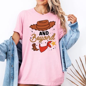 Peut inclure: T-shirt rose avec une image de dessin anim&eacute; du chapeau de Woody de Toy Story. Le t-shirt porte le texte "AND Beyond" en lettres dor&eacute;es scintillantes.