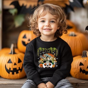 May include: A child wearing a black sweatshirt with a Halloween-themed graphic. The graphic features various cartoon animals, pumpkins, and bats. The background includes several carved orange pumpkins.