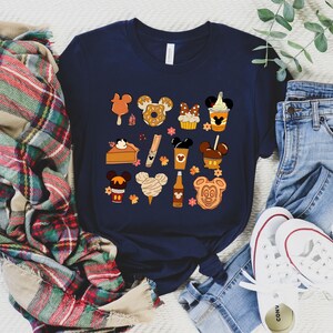 May include: Navy blue t-shirt featuring a collection of cartoon food items with Mickey Mouse ears. The illustrations include a popsicle, donut, cupcake, pie, churro, coffee, caramel apple, cotton candy, and a Mickey Mouse-shaped cookie.