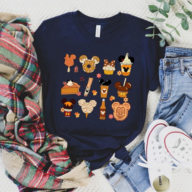 May include: Navy blue t-shirt featuring a collection of cartoon food items with Mickey Mouse ears. The illustrations include a popsicle, donut, cupcake, pie, churro, coffee, caramel apple, cotton candy, and a Mickey Mouse-shaped cookie.