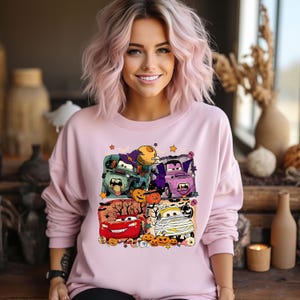 May include: Pink sweatshirt with a Halloween-themed graphic featuring cartoon cars dressed up as a witch, vampire, mummy, and a red car with a pumpkin on top. The graphic includes pumpkins, bats, and candy corn.