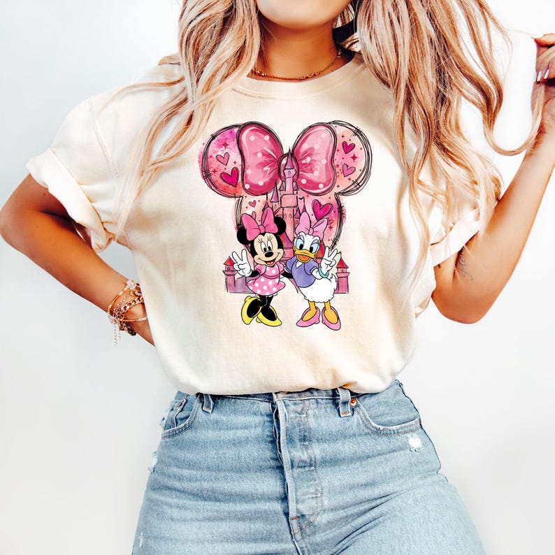 May include: Cream-colored t-shirt featuring a graphic of Minnie Mouse and Daisy Duck. The design includes a pink bow, hearts, and a castle. Minnie Mouse wears a pink polka-dot dress, and Daisy Duck is in a purple top.
