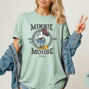 May include: A light green t-shirt featuring a vintage-style graphic of Minnie Mouse. The design includes the text "MINNIE MOUSE" and the year "1928" in a circular frame. The shirt is part of the Comfort Colors Bay collection.