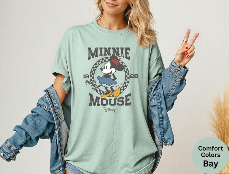 May include: A light green t-shirt featuring a vintage-style graphic of Minnie Mouse. The design includes the text "MINNIE MOUSE" and the year "1928" in a circular frame. The shirt is part of the Comfort Colors Bay collection.