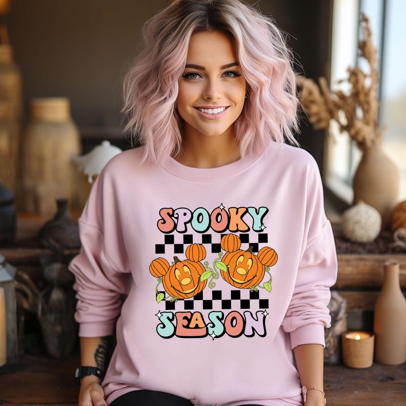 May include: A light pink sweatshirt with the words "Spooky Season" in a retro font. The design includes two orange jack-o'-lanterns with Mickey Mouse ears on a black and white checkered background. The sweatshirt is a crew neck style.