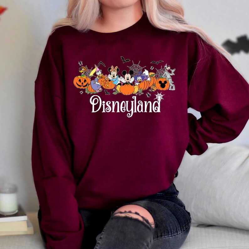 K&ouml;nnte beinhalten: Ein weinrotes Sweatshirt mit einem Halloween-Design von Disneyland. Die Grafik zeigt Zeichentrickfiguren, K&uuml;rbisse, Flederm&auml;use und Spinnweben. Das Wort "Disneyland" ist in wei&szlig;er Schrift geschrieben.