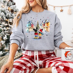 Op de afbeelding: Een grijze sweatshirt met een winterse sc&egrave;ne met Mickey en Minnie Mouse. De afbeelding toont het paar voor kleurrijke gebouwen en een kerstboom. De sweatshirt wordt gecombineerd met een rood-witte geruite pyjamabroek.