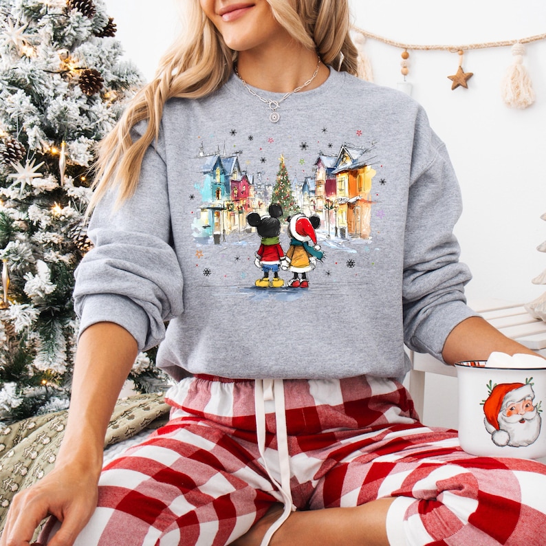 Op de afbeelding: Een grijze sweatshirt met een winterse sc&egrave;ne met Mickey en Minnie Mouse. De afbeelding toont het paar voor kleurrijke gebouwen en een kerstboom. De sweatshirt wordt gecombineerd met een rood-witte geruite pyjamabroek.
