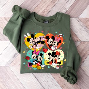 May include: Green sweatshirt with a graphic print of Mickey Mouse and Minnie Mouse in four different poses. Each pose is inside a heart shape with a different color background. The hearts are surrounded by small pink hearts.