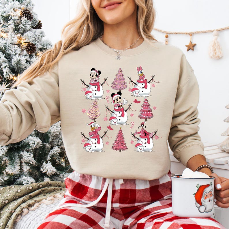 May include: Beige crew neck sweatshirt with a repeating pattern of snowmen with Disney characters, pink Christmas trees, and candy canes. The snowmen wear hats and scarves. A Santa mug is held in the lower right corner.