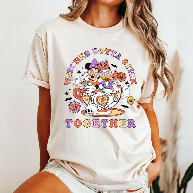 K&ouml;nnte beinhalten: Ein beiges T-Shirt mit einem Aufdruck von Mickey Mouse und Daisy Duck, die als Hexen in einer Teetasse verkleidet sind. Der Text "Witches Gotta Stick Together" ist auf dem Shirt aufgedruckt.
