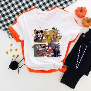 Puede incluir: Camiseta blanca con un dise&ntilde;o de Halloween que presenta a Mickey Mouse, Goofy y Donald Duck disfrazados. La camiseta tiene un ribete naranja. La imagen incluye pantalones negros y decoraciones de Halloween.