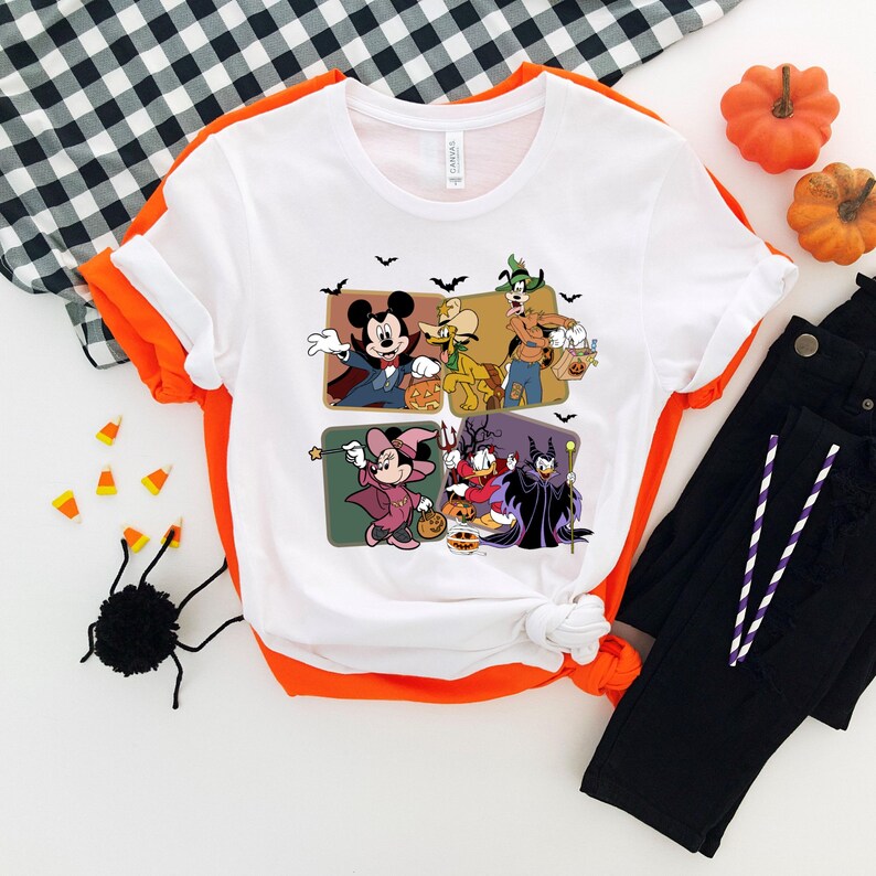 Puede incluir: Camiseta blanca con un dise&ntilde;o de Halloween que presenta a Mickey Mouse, Goofy y Donald Duck disfrazados. La camiseta tiene un ribete naranja. La imagen incluye pantalones negros y decoraciones de Halloween.