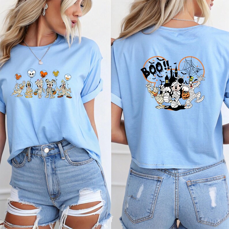 K&ouml;nnte beinhalten: Hellblaues T-Shirt mit Halloween-Motiven. Auf der Vorderseite sind Cartoon-Figuren mit Luftballons abgebildet. Auf der R&uuml;ckseite steht das Wort "BOO!!" und ein Geisterhaus. Mit hochgekrempelten &Auml;rmeln.