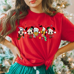 May include: Red Christmas themed t-shirt featuring Mickey Mouse, Minnie Mouse, Donald Duck, Goofy, and Pluto wearing Santa hats and scarves.