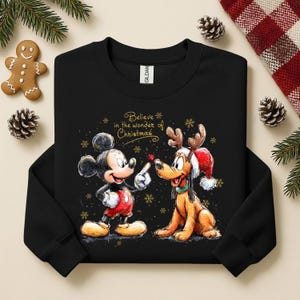 May include: Black sweatshirt featuring Mickey Mouse and Pluto with Christmas-themed designs. Mickey Mouse is wearing red shorts and Pluto is wearing a Santa hat. The text "Believe in the wonder of Christmas" is displayed in gold.