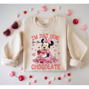 May include: Cream-colored sweatshirt with a Minnie Mouse graphic holding a heart-shaped gift box. The text "I'M JUST HERE for the CHOCOLATE" is printed in red. Pink heart-shaped sunglasses and candies surround the sweatshirt.