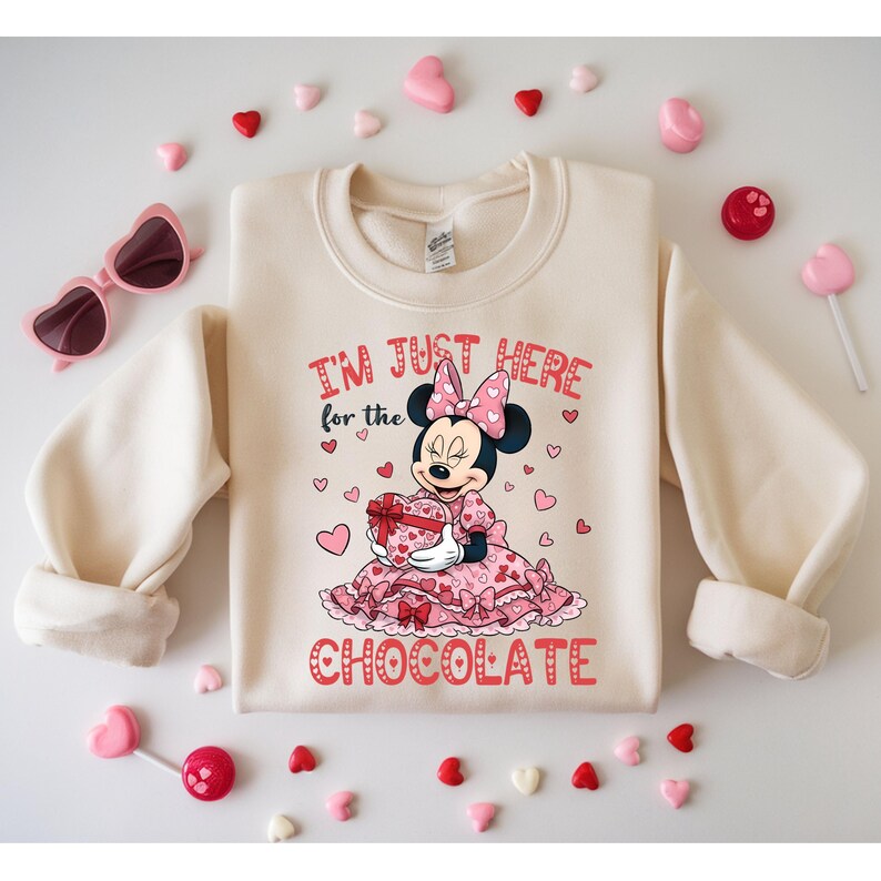 May include: Cream-colored sweatshirt with a Minnie Mouse graphic holding a heart-shaped gift box. The text "I'M JUST HERE for the CHOCOLATE" is printed in red. Pink heart-shaped sunglasses and candies surround the sweatshirt.