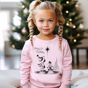 May include: A light pink sweatshirt featuring a graphic of a cartoon character and a dog, with a castle in the background. The text "Believe in the wonder of Christmas" is printed above the graphic. The sweatshirt is worn by a young girl with blonde braids.