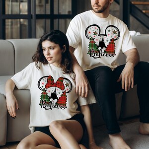 Puede incluir: Una pareja lleva camisetas blancas con un dise&ntilde;o navide&ntilde;o con orejas de Mickey Mouse, un lazo de cuadros, una silueta del castillo de Cenicienta y el texto "Believe".