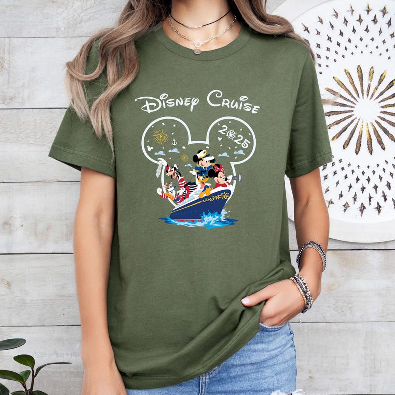 May include: Olive green t-shirt featuring the words "Disney Cruise" above a Mickey Mouse head outline. Inside the outline is a cruise ship with Mickey, Minnie, Donald, and Goofy. The year "2025" is also inside the outline.