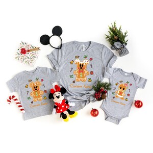 May include: A set of three grey t-shirts featuring a gingerbread Mickey Mouse design. The shirts are available in adult and child sizes. The shirts have the text "Custom Name" printed on them.